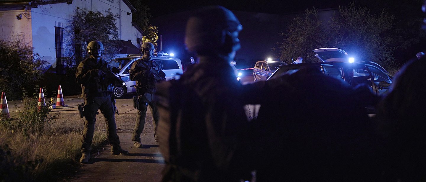 Polizeibeamte mit schwerer Ausrüstung sind im Dunkeln vor einem Haus unterwergs. 