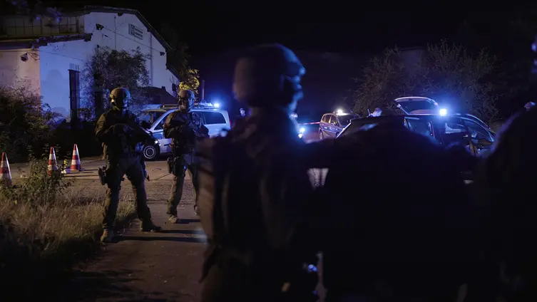 Polizeibeamte mit schwerer Ausrüstung sind im Dunkeln vor einem Haus unterwergs. 