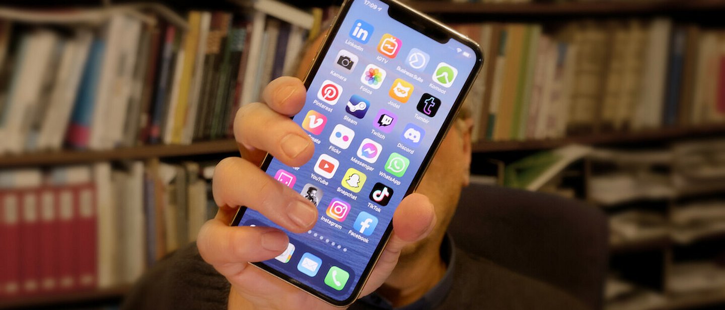 Man sieht ein Handy/IPhone das von einer Hand mit den verschiedensten Apps für Socialmedia in richtung der Kamera gehalten wird. Im hintergrund sieht man unscharf ein Bücherregal und die Person die das Handy hält.