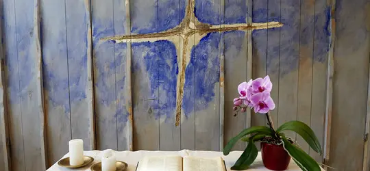 Holzwand mit blau gemaltem Kreuz. Auf einem Tisch davor stehen zwei weiße Kerzen, ein aufgeschlagenes Buch und eine rosa Orchidee im Topf.
