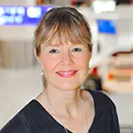 Porträt einer lächelnden Frau mit blondem Haar und Pony. Sie trägt ein dunkles Oberteil und befindet sich in einem hellen Flughafenumfeld mit unscharfem Hintergrund. Das Bild zeigt Frau Bach.