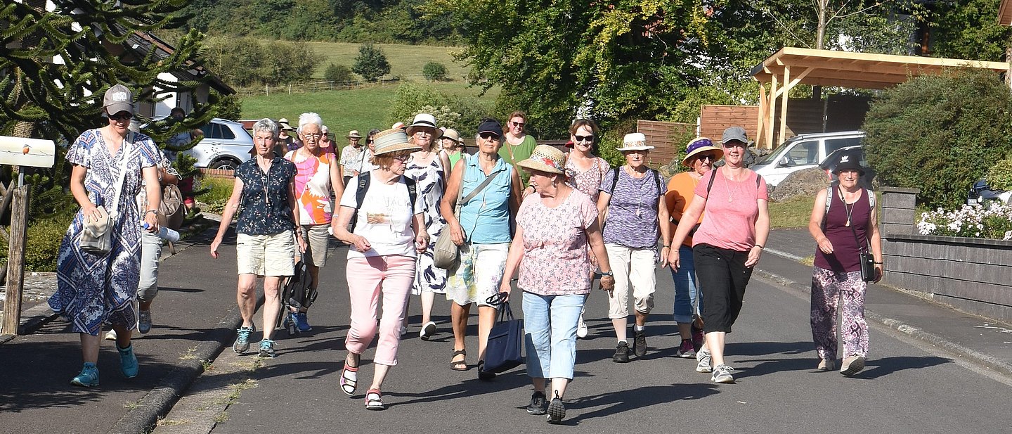 Eine große Gruppe Frauen, sommerlicher Freizeitlook, zum Teil mit Hüten, läuft eine Dorfstraße entland