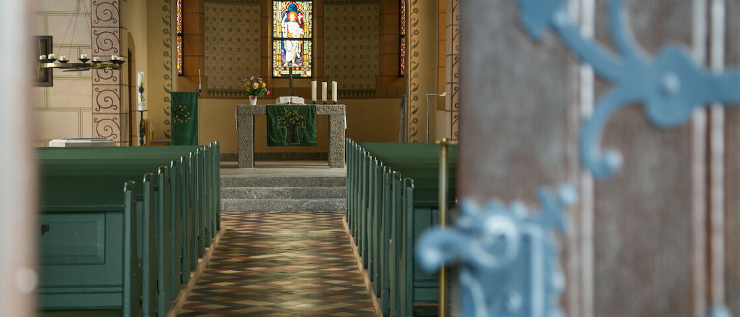 Im Vordergrund rechts die Eingangstür zur Kirche. Bunte Bodenfliesen, Verzierungen am Bogen. Helle Fenster, im Altarraum hellgrauer Steinboden.