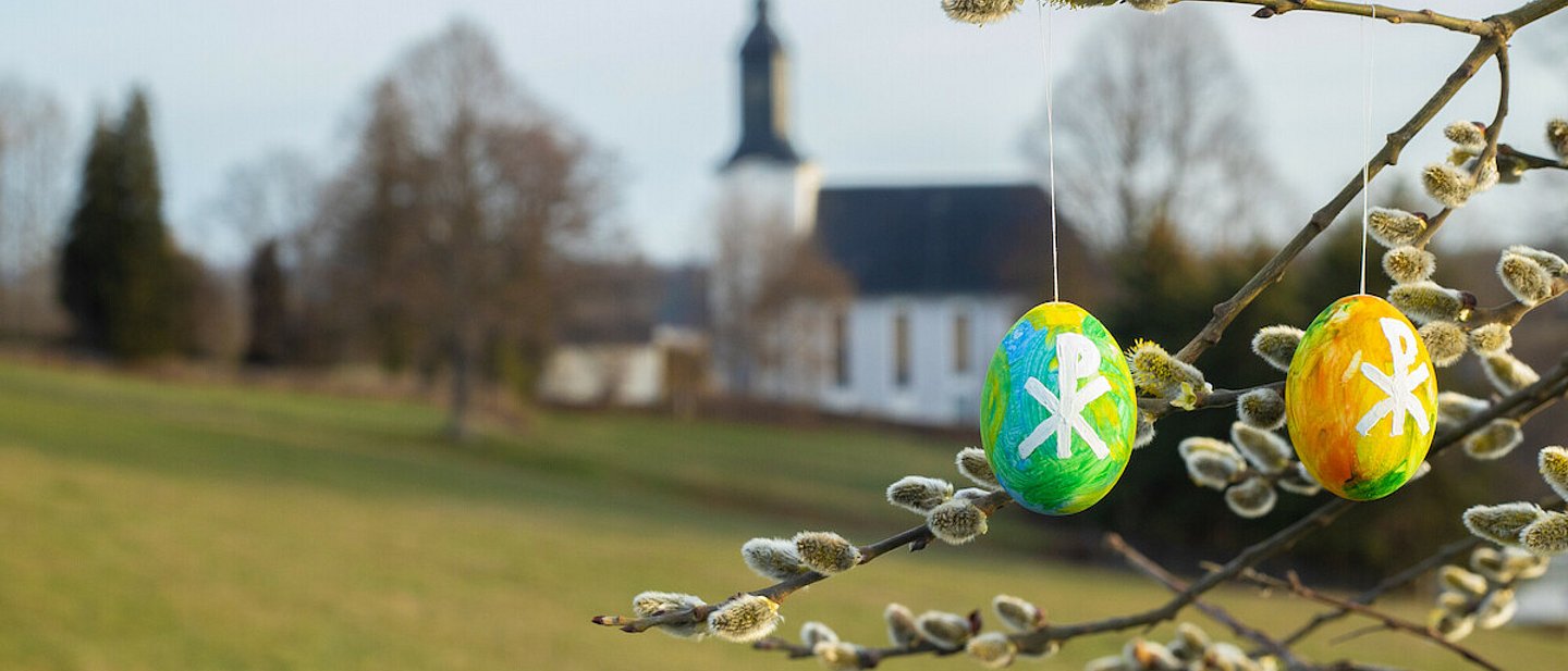 Bunt bemalte Ostereier hängen an einem Baum. Im Hintergrund ist eine Dorfkirche zu sehen.