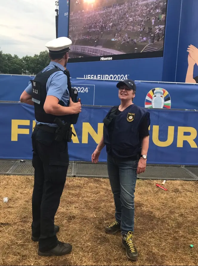 Die Pfarrerin steht rechts, der Polizist ihr zugewandt steht links. Beide befinden sich in der Fanzone der UEFA-Europameisterschaft in Frankfurt. Im Hintergrund ein riesiger Bildschirm.
