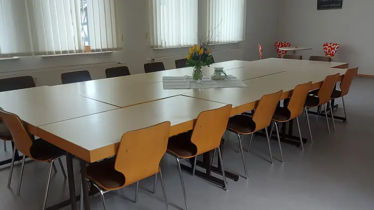 Ein heller Raum. In der Mitte steht ein großer Tisch, um den Tisch herum stehen Stühle