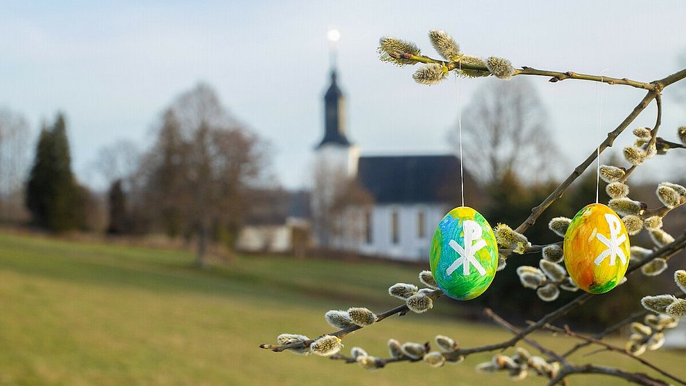 Ostereier Bunt bemalte Ostereier hängen an einem Baum. Im Hintergrund ist eine Dorfkirche zu sehen.