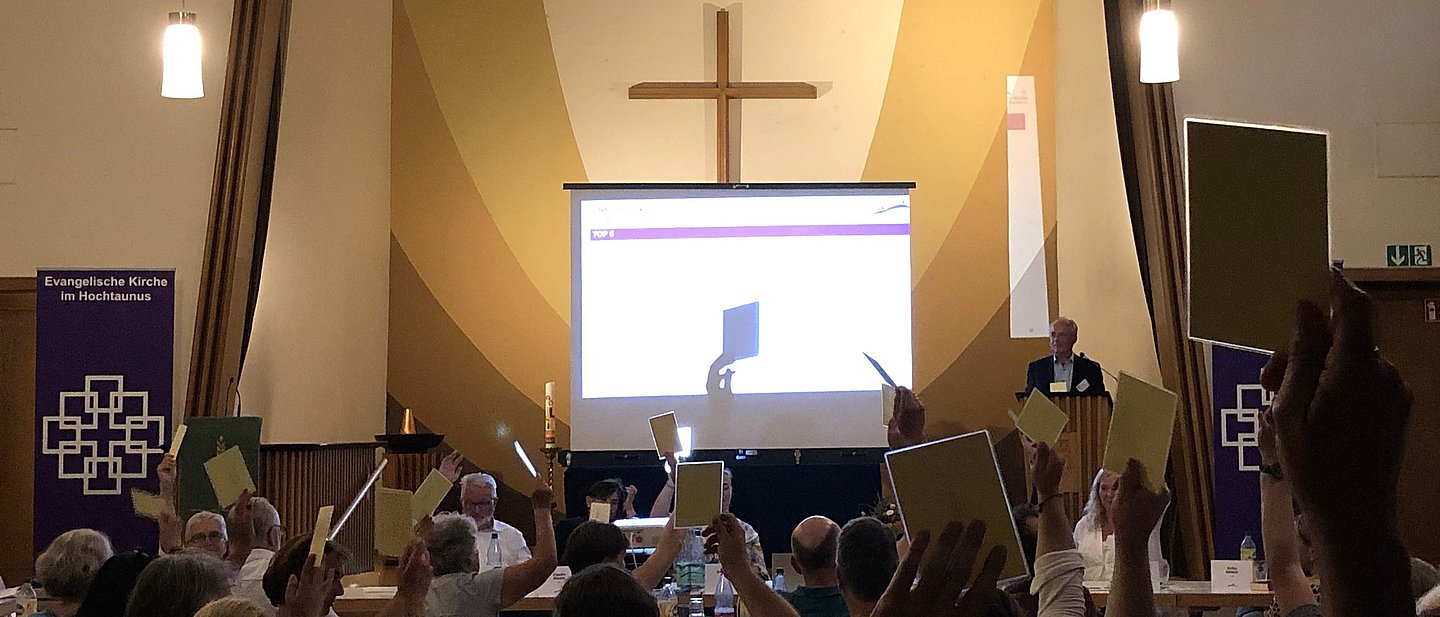 Tagung einer Synode in einem modernen Kirchenraum. Menschen heben die Hände mit Abstimmungskarten. Im Hintergrund eine Projektion mit Beamer. 