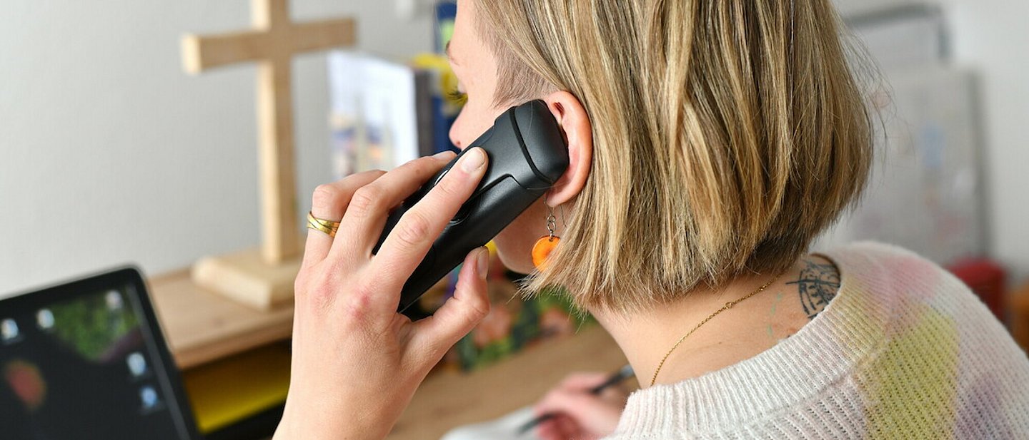 Pfarrerin Nele Schomakers in einem Büro beim Telefonieren - seitlich von hinten aufgenommen mit Blick auf den Bürotisch.  In der linken Hand das Telefon, in der rechten Hand ein Kugelschreiber um Notizen zu machen. Im Vordergrund die Hand und der Telefonhörer am Ohr.