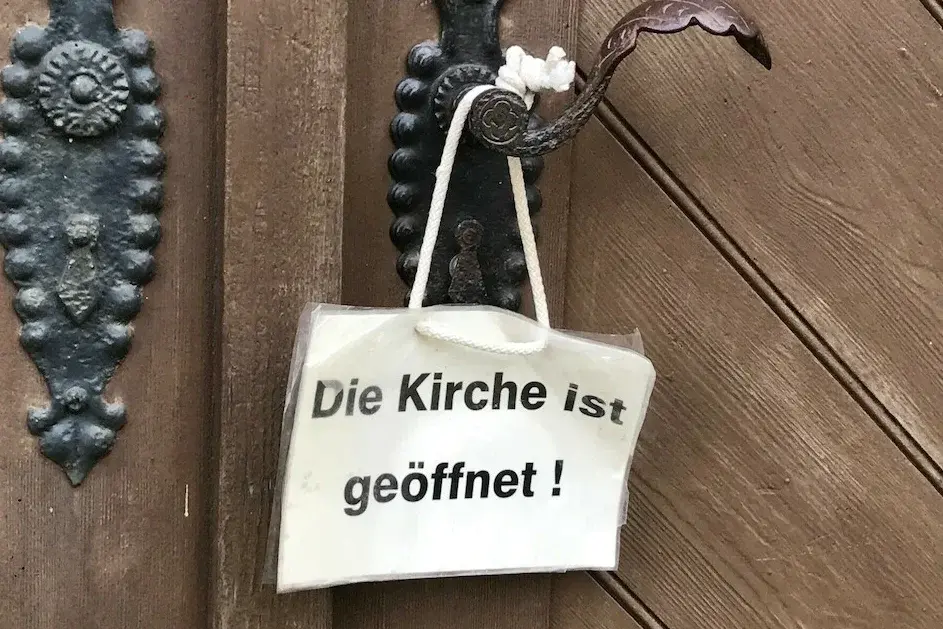 Weißes Schild hängt an Klinke der Kirchentür. Auf dem Schild steht: "Die Kirche ist geöffnet".