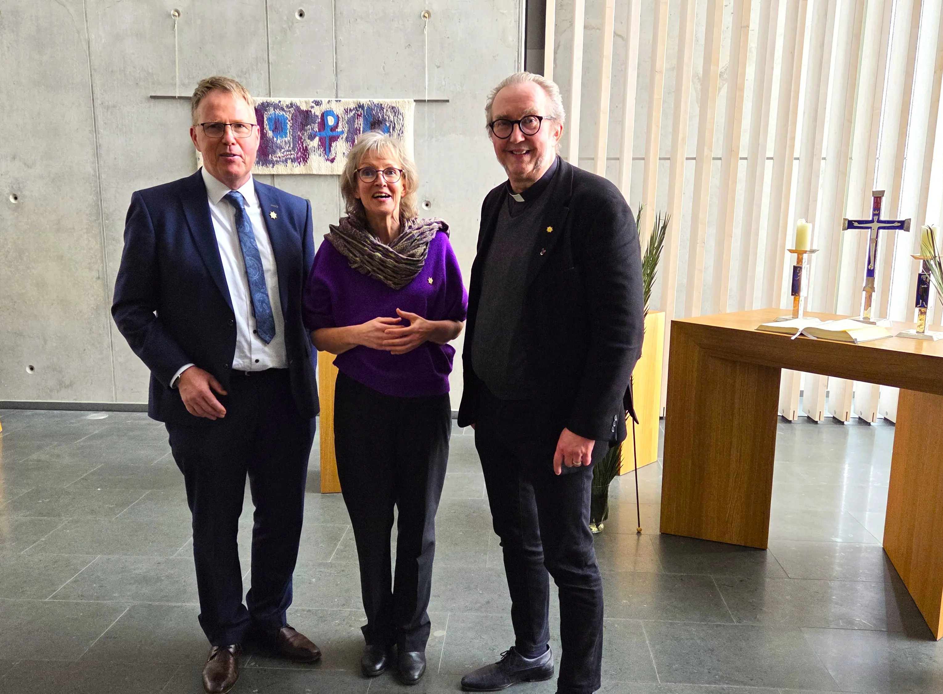 Die drei Personen stehen nebeneinander in einem Kirchenraum vor einem Altar
