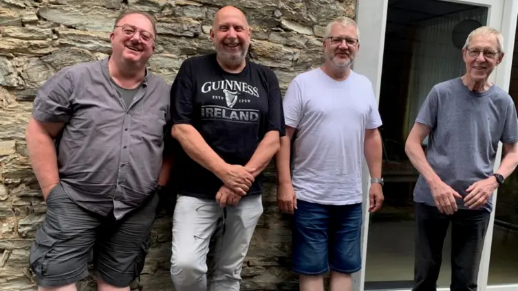 Das Bild zeigt eine Gruppe von vier Männern, die vor einem Gebäude stehen. Sie sind alle mittleren Alters oder älter zu sein und sind leger gekleidet. Im Hintergrund ist eine Steinmauer und eine Tür oder ein Fenster zu sehen. Die Männer scheinen gut gelaunt zu sein.