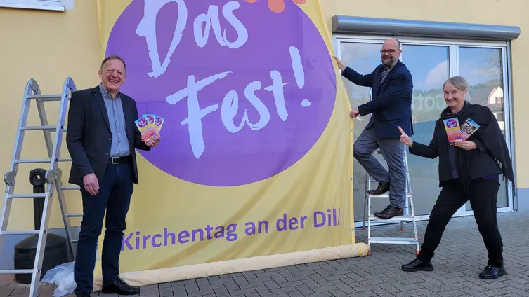 Mit dem Super-Banner „Das Fest!“ wirbt das Evangelische Dekanat an der Dill am Herborner „Haus der Kirche und Diakonie“ für den Dill-Kirchentag am 12. Und 13. September 2026. Präses Dr. Wolfgang Wörner (von links), Dekan Andree Best und die stellvertretende Dekanin Anja Vollendorf freuen sich über den Austausch und die Begegnung.