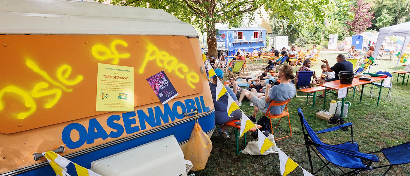 Man sieht im Vordergrund das Oasenmobil und im Hintergrund Jugendliche auf Bänken und Stühlen