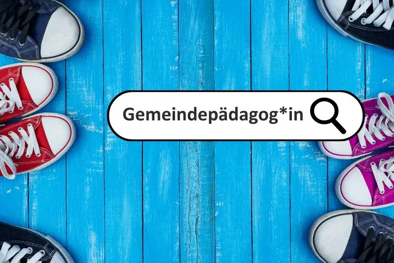 Ein Suchfeld mit dem Wort "Gemeindepädagog*in" und dem Ikon einer Lupe vor einem Hintergrund aus blau gestrichenen Holzbalken. Am rechten Bildrand sind die Anschnitte von Sneakers zu sehen. 