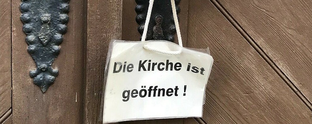 Weißes Schild hängt an Klinke der Kirchentür. Auf dem Schild steht: "Die Kirche ist geöffnet".