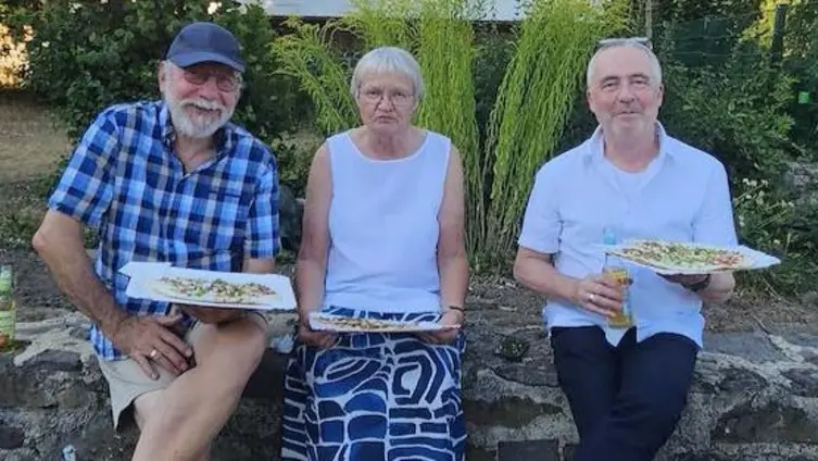 Zwei Männer, eine Frau, sitzen auf einer Basaltsteinmauer, hinter sich das Niddaer Schloss, und halten Flammkuchen in Händen. Alle tragen legere Freizeitkleidung.