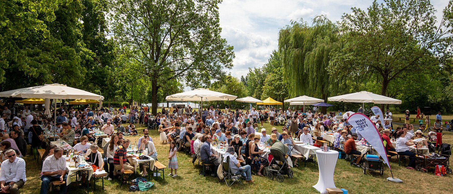 Hunderte Leute sitzen bei Sonnenschein unter Sonnenschirmen auf Bierbänken und feiern das "Main Tauffest 2022" am Mainufer in Offenbach.