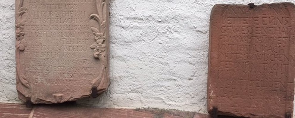 Zwei Sandstein-Grabplatten an der Wand der evangelischen Kirche Köppern. Die linke mit floralem Randschmuck von 1753. Die rechte, kleinere, aus dunkleren Sandstein von 1615.