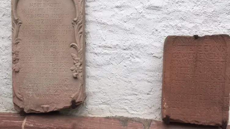 Zwei Sandstein-Grabplatten an der Wand der evangelischen Kirche Köppern. Die linke mit floralem Randschmuck von 1753. Die rechte, kleinere, aus dunkleren Sandstein von 1615.