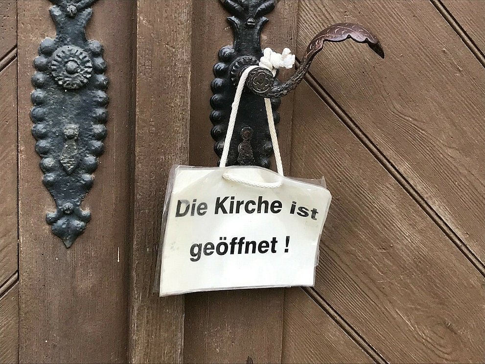 Weißes Schild hängt an Klinke der Kirchentür. Auf dem Schild steht: "Die Kirche ist geöffnet".