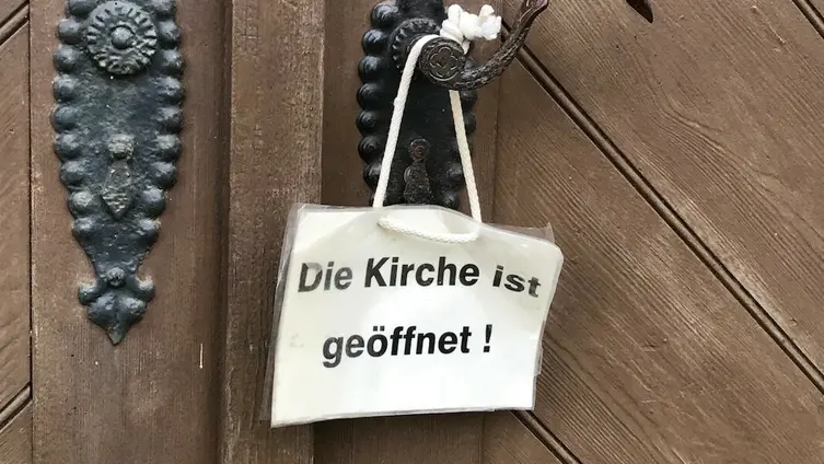 Weißes Schild hängt an Klinke der Kirchentür. Auf dem Schild steht: "Die Kirche ist geöffnet".