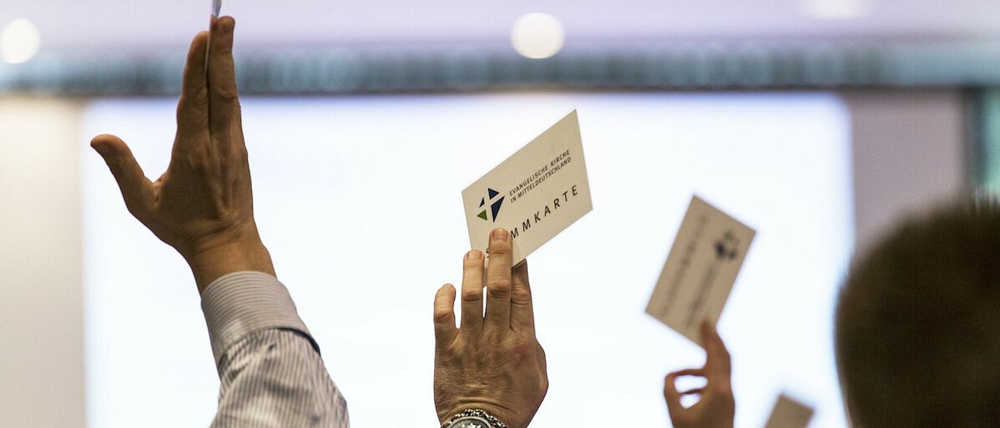 Man sieht drei in die Luft gestreckte Hände. Sie halten Stimmkarten mit der Aufschrifft "Stimmkarte - Evangelische Kirche Mitteldeutschland" hocht.
