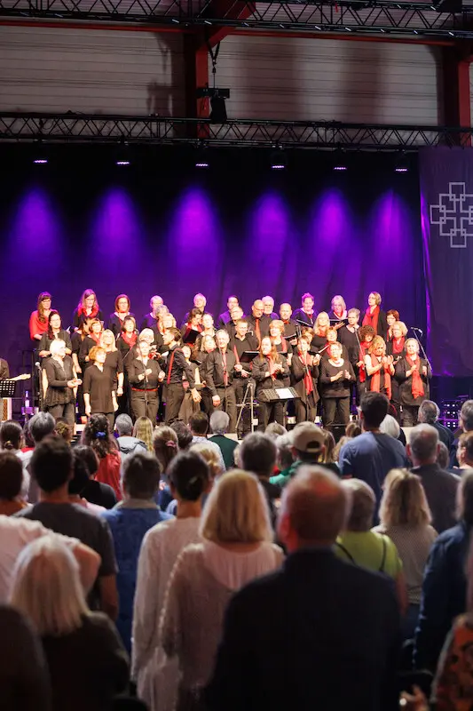 Ideenmesse der EKHN in Gießen. Blick über das von hinten aufgenommenen Publikum auf die Bühne mit dem Chor und einem Pfarrer. Das Publikum streckt die Arme in die Höhe. Hochformat.