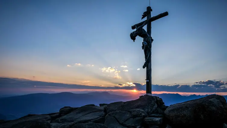 Man sieht einen Berggipfel mit dem dem überlebensgroßen Christus am Kreuz vor einem Abendhimmel mit Sonnenuntergang