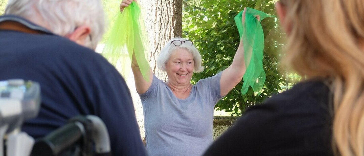Dorothee Ewald steht strahlend vor Senior*innen und hält zwei grüne Tücher in die Höhe. 