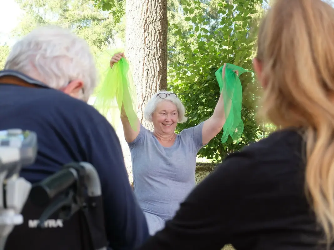 Dorothee Ewald steht strahlend vor Senior*innen und hält zwei grüne Tücher in die Höhe. 