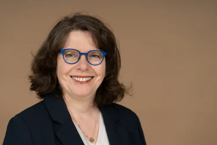 Porträt Kerstin Hillgärtner, dunkle, schulterlange Locken, Brille, freundliches Lachen, vor einem neutralen Hintergrund