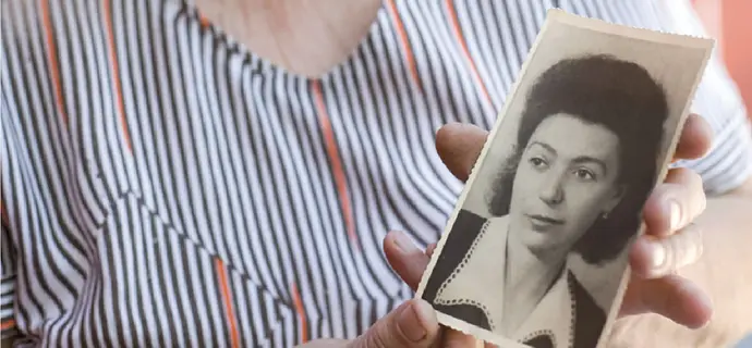 Bildausschnitt einer Seniorin, die ein altes schwarz/weiß-Portrait-Foto von einer jungen Frau in die Kamera hält.