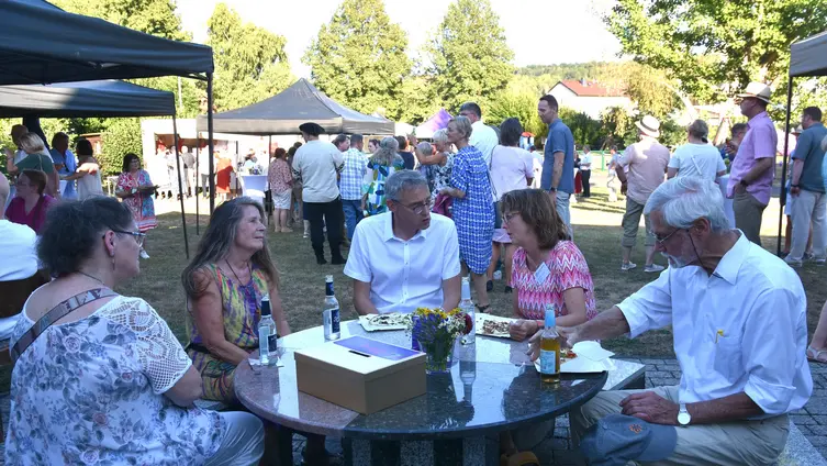 Blick in den Pfarrgarten, im Vordergrund Menschen an einer fest installierten Sitzgruppe aus Stein, dahintere sieht man das Gelände mit Stehtischen und Ständen.