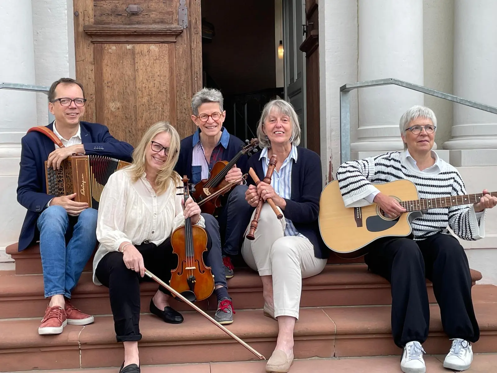 5 Menschen sitzen vor der offenen Kirchentür. Links der Musiker hat ein kleines Akkordeon auf dem Schoß, dann sind zwei Damen mit Violine zu sehen, daneben sitzt eine Frau mit Flöten in der Hand, ganz rechts sitzt eine weißhaarige Frau mit einer Gitarre. Es ist die Taunussteiner Folkband