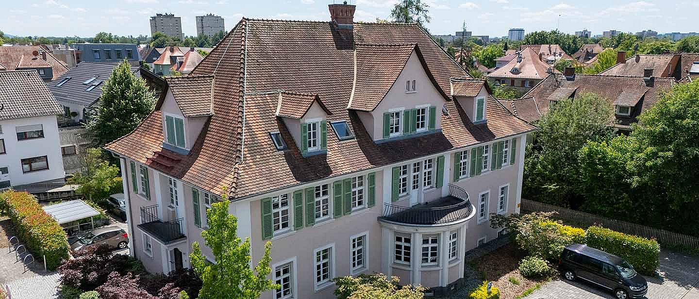 Großes mehrstöckiges Gebäude mit rotem Ziegeldach, hellen Fassaden und grünen Fensterläden, umgeben von Bäumen, Gärten und parkenden Autos.