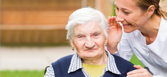 Eine ältere Frau mit weißem Haar sitzt im Freien und schaut freundlich in die Kamera. Neben ihr beugt sich eine jüngere Pflegekraft zu ihr hin und hält eine Hand ans Ohr der Seniorin, um ihr beim Hören zu helfen. Beide wirken entspannt und zugewandt. Eine ältere Frau mit weißem Haar sitzt im Freien und schaut freundlich in die Kamera. Neben ihr beugt sich eine jüngere Pflegekraft zu ihr hin und hält eine Hand ans Ohr der Seniorin, um ihr beim Hören zu helfen. Beide wirken entspannt und zugewandt.