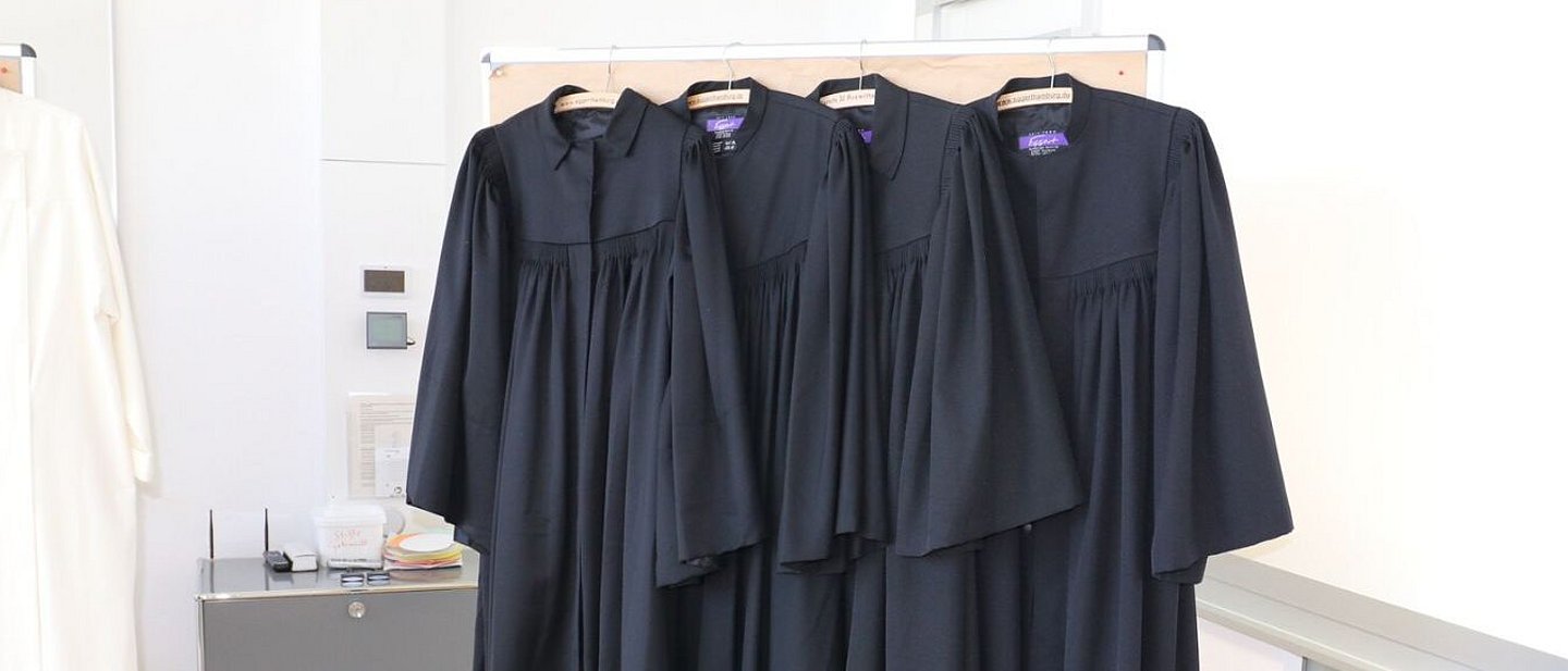 vier schwarze Talare hängen auf Kleiderbügeln an einer Moderationswand