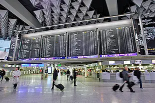 Große Abfluganzeigetafeln in einem modernen Flughafenterminal. Reisende laufen mit Koffern durch die helle Halle, während im Hintergrund Check-in-Schalter und Informationsanzeigen zu sehen sind.