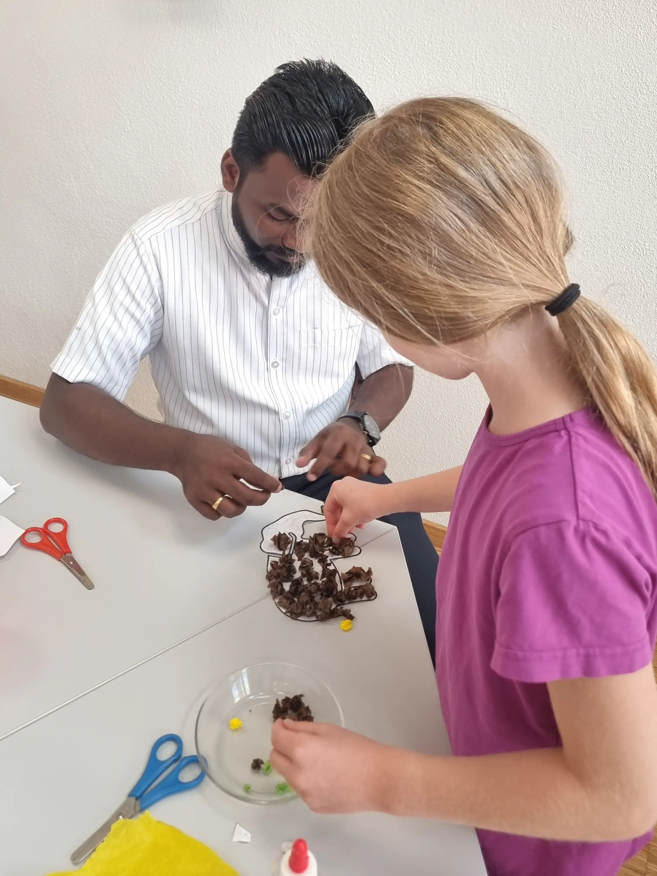 Mann und Mädchen sitzen am Tisch und basteln. Vor ihnen liegen Scheren, Kleber, Papier und Stifte.