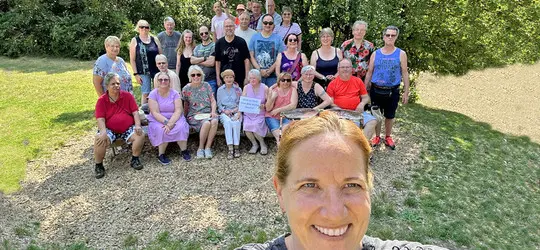 Frau macht ein Selfie vor einer großen Gruppe lachender Menschen im Freien bei sonnigem Wetter.