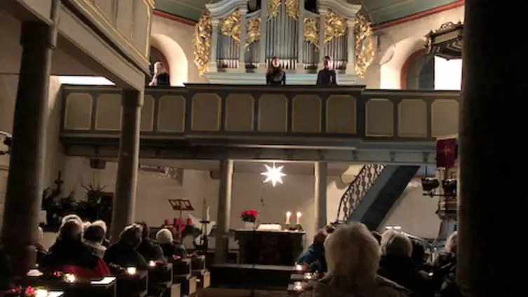 Blick vom Haupteingang der evangelischen Kirche Köppern in den nur mit Kerzen erhellten Besucherraum. Über dem Altar hängt der erleuchtete Herrnhuter Stern. Die Orgelempore darüber ist beleuchtet.
