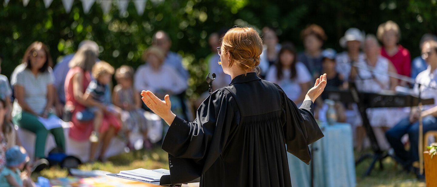 Tauffest im Garten der Pfingstkirche in Frankfurt-Griesheim am 25.06.2023. Im Vordergrund steht die Pfarrerin mit dem Rücken zur Kamera. Dahinter im unscharfen Bereich die Besucher*innen der Taufveranstaltung.