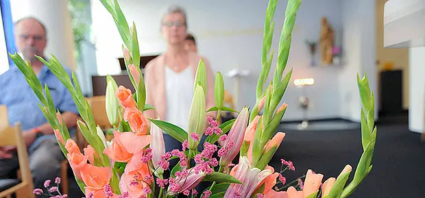 Großer Blumenstrauß mit rosa und orangefarbenen Blüten im Vordergrund eines Andachtsraums. Im unscharfen Hintergrund sitzen zwei Personen auf Stühlen, Kerzenlicht und religiöse Symbole schaffen eine ruhige Atmosphäre.