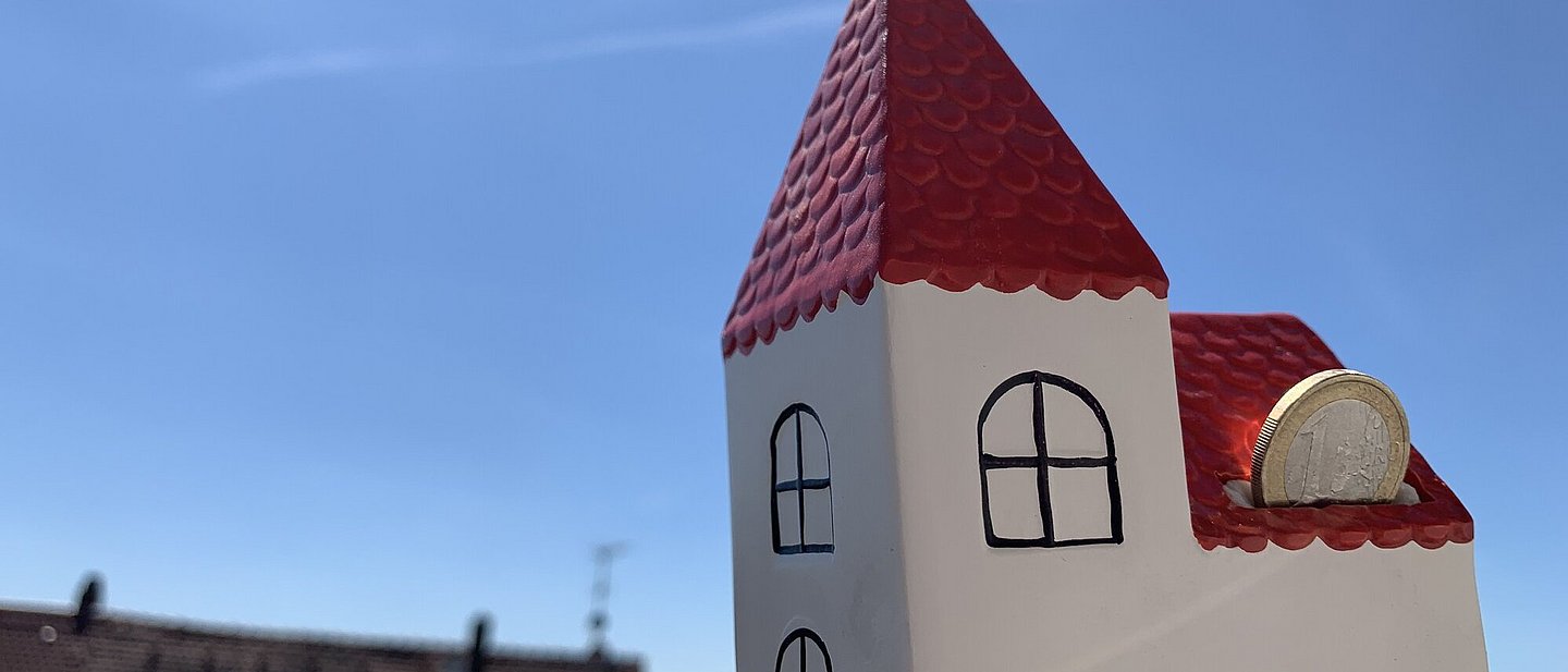 Spardose in Form einer weißen Kirche mit rotem Dach. Eine 1 € -Münze wird gerade durch den Schlitz geschoben