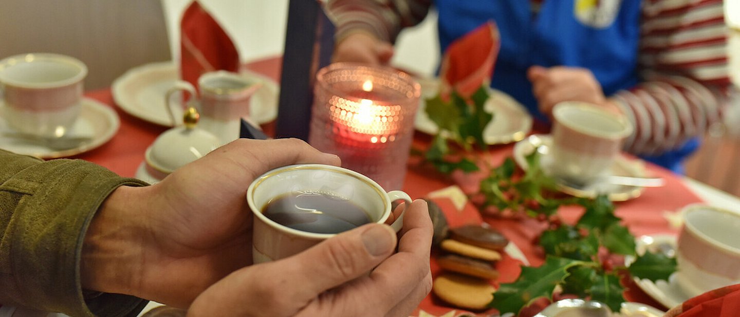Zu sehen sind zwei Personen, die gerade an einem gedeckten Tisch sitzen und Kaffee trinken und dabei Gebäck essen.