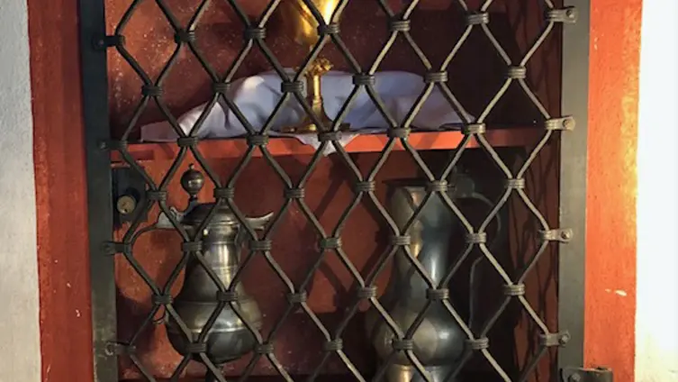 Die in der linken Chorwand der evangelischen Kirche Köppern eingelassene, sandsteinrot gefasste Abendmahlsnische mit Gitter davor. Darin auf dem Regalbrett oben ein goldener Kelch. Darunter zwei Weinkannen aus Zinn. Die linke aus dem Barock, bauchig, auf einem Fuß stehend. 