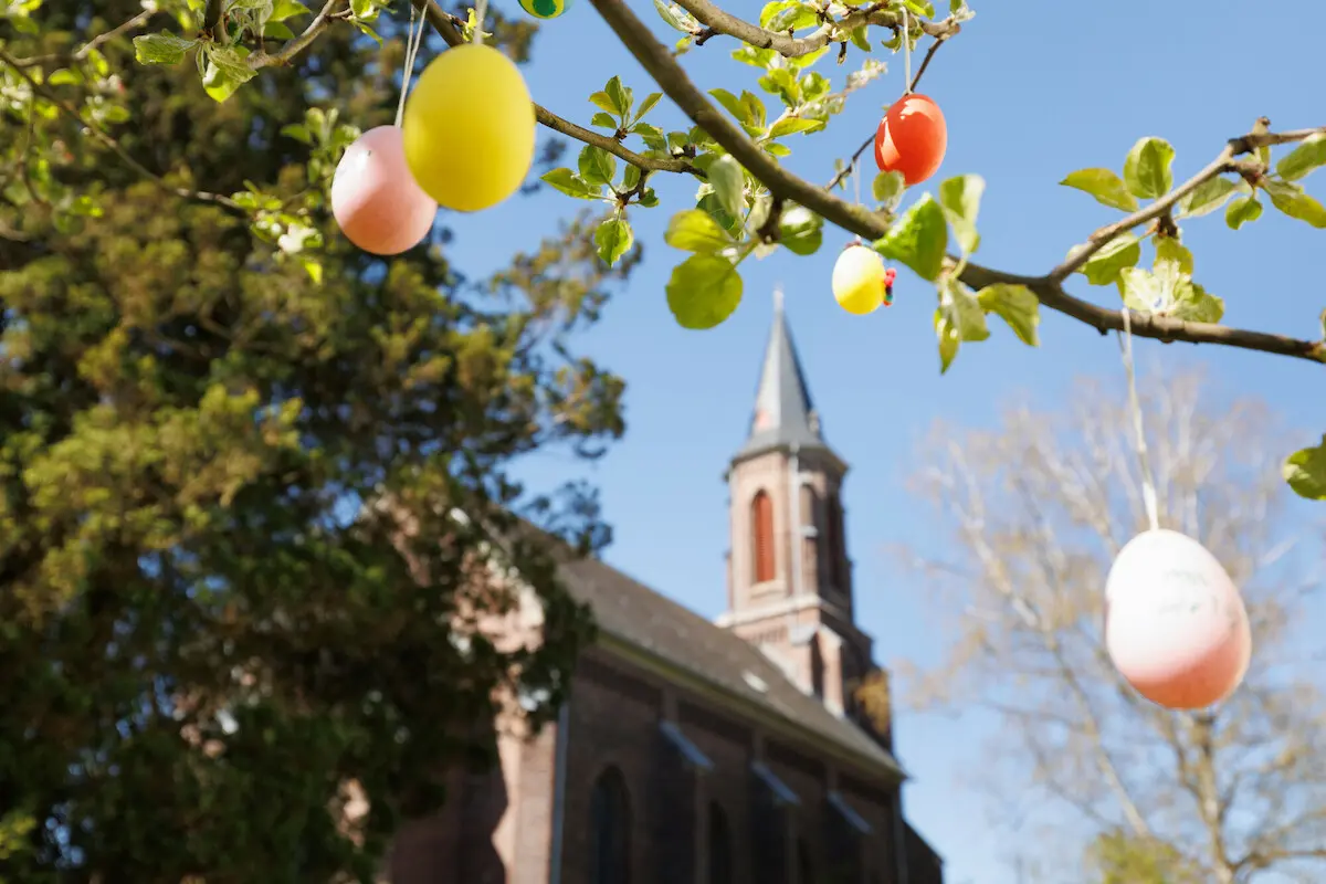 Blick auf einen Kirchturm unter blauem Himmel, davor Zweige eines vermutlich Obstbaumes, der mit bunten Ostereiern geschmückt ist.