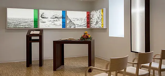 Schlichter Andachtsraum mit Tisch, Lesepult, Blumenstrauß und Wandkunst in mehreren Farbakzenten.