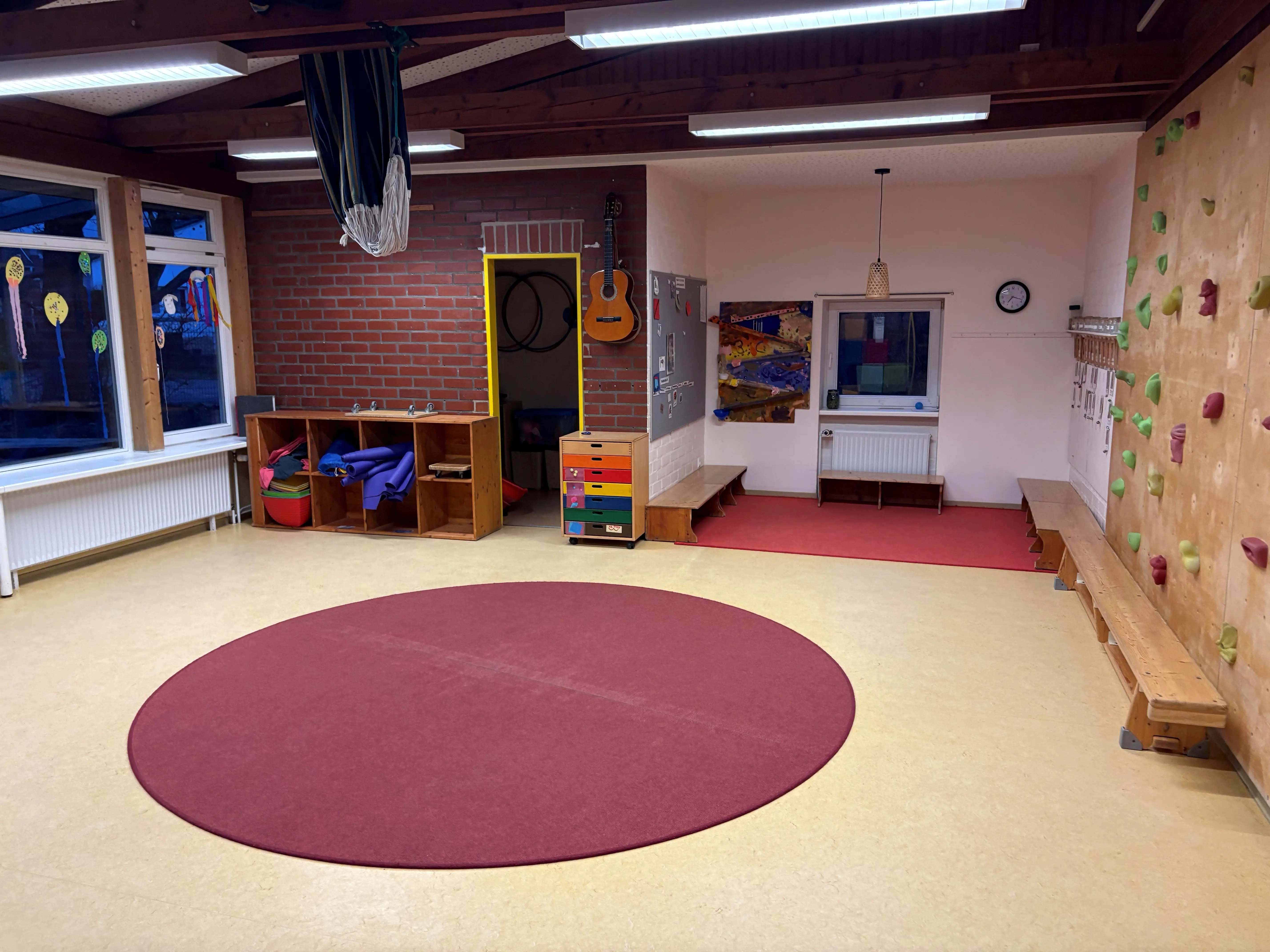 Bild unseres Bewegungsraums,ein roter runder Teppich liegt in der Mitte, an der rechten Wand ist eine Kletterwand mit roten grünen und gelben Klettergriffenn zu sehen. geradeaus stehen kleine und eine große Bank. Links im Bild ein Regal mit Flusssteinen Tüchern, Yogamatten und einem Rollbrett. Daneben eine Tür, welche zu unserem Matarialraum führt, an der Wand sind Reifen zu erkennen.